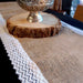 Burlap table runner with white lace trim edge displayed on a wooden surface next to an ornate brass pedestal bowl.