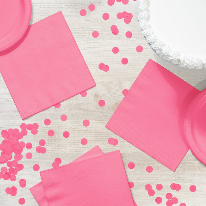 Bright pink paper napkins arranged on a white wooden surface with scattered pink confetti circles and a white frosted cake visible in the corner.