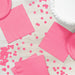 Bright pink paper napkins arranged on a white wooden surface with scattered pink confetti circles and a white frosted cake visible in the corner.