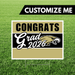 Customizable yard sign with gold and black horizontal stripes, white text reading "CONGRATS Grad 2026" with an eagle mascot logo, displayed on green grass.