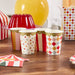 Four decorated paper cups with gold scalloped rims sit on a wooden surface among circus-themed party decorations including red and white striped items and a yellow balloon.