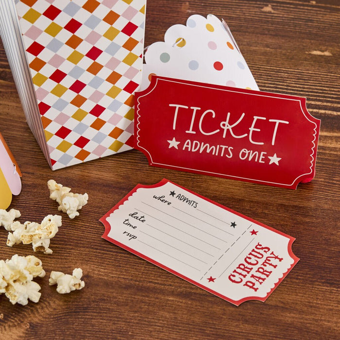 Red ticket-shaped invitations with "Ticket Admits One" text displayed with popcorn, a colorful diamond-patterned paper bag, and a white polka-dotted envelope on a wooden surface.