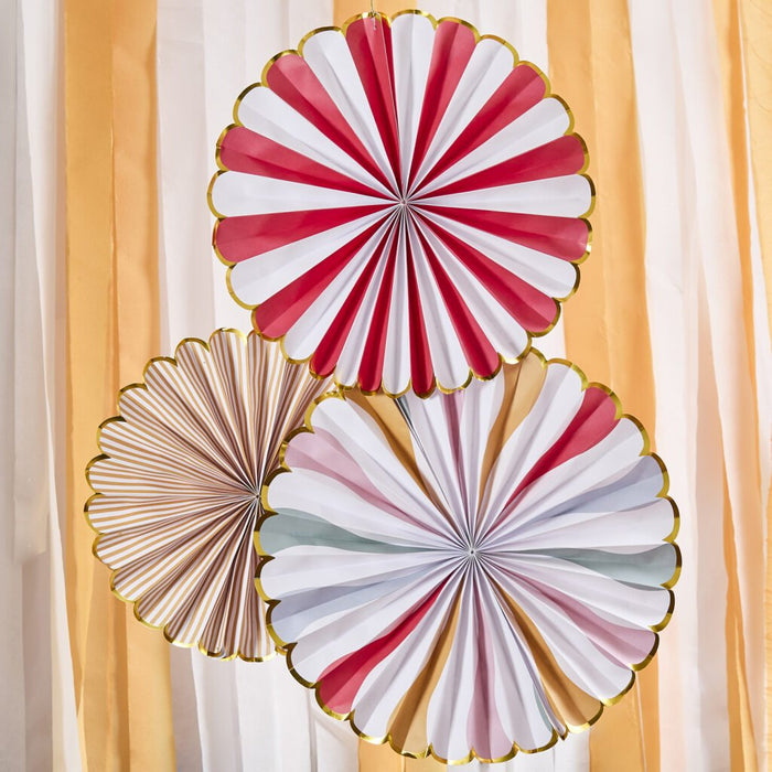 Three paper fan decorations with scalloped edges in red and white stripes, tan stripes, and multicolor pastels, displayed against a striped yellow and white backdrop.