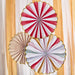 Three paper fan decorations with scalloped edges in red and white stripes, tan stripes, and multicolor pastels, displayed against a striped yellow and white backdrop.