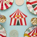 Red and white striped circus tent napkins with a gold "Circus" label, surrounded by coordinating striped plates, popcorn containers, and pastel swirl decorations on a light blue background.