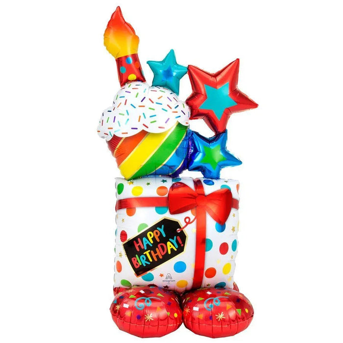 Tall colorful stacked birthday balloon arrangement with rainbow ice cream cone, lit candle, stars, gift box with red bow, and red base balloons, featuring "Happy Birthday" text on polka-dotted white section.