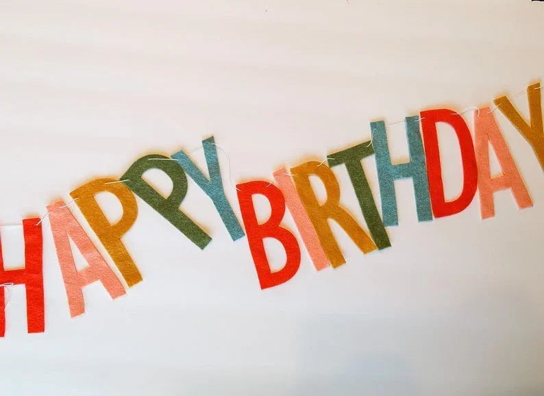Colorful felt banner strung across white background with staggered letters spelling "Happy Birthday" in red, orange, yellow, green, and blue.