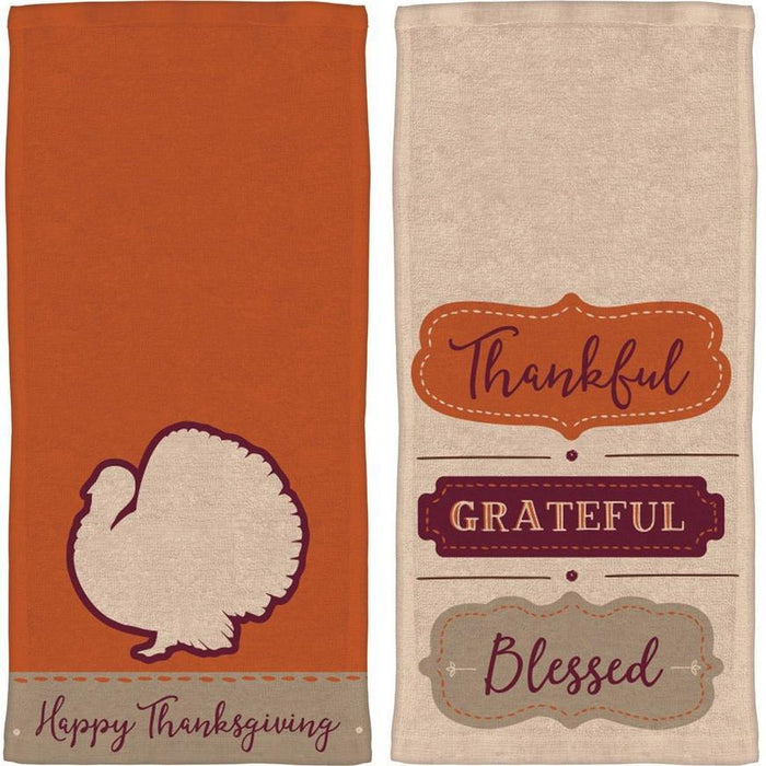 Two Thanksgiving-themed cotton dish towels: one orange with a cream turkey silhouette and "Happy Thanksgiving" text, and one cream with decorative labels reading "Thankful," "Grateful," and "Blessed" in burgundy and brown.