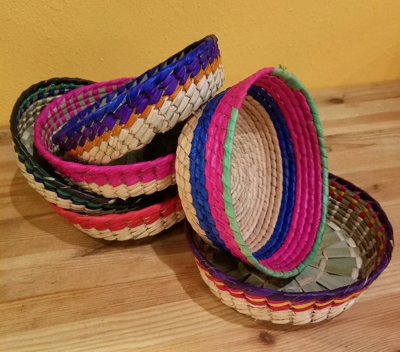 Four colorful woven straw baskets with bright pink, blue, green, purple, and natural tan striped trim arranged on a wooden surface.