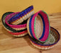 Four colorful woven straw baskets with bright pink, blue, green, purple, and natural tan striped trim arranged on a wooden surface.