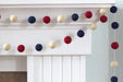 Red, navy, and cream felt ball garland strung horizontally across a white mantelpiece and draped vertically down the side in a patriotic color pattern.