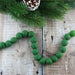 Green felt ball garland with approximately 24 pompoms arranged in a U-shape, displayed on weathered wood with an evergreen branch and pinecone above.