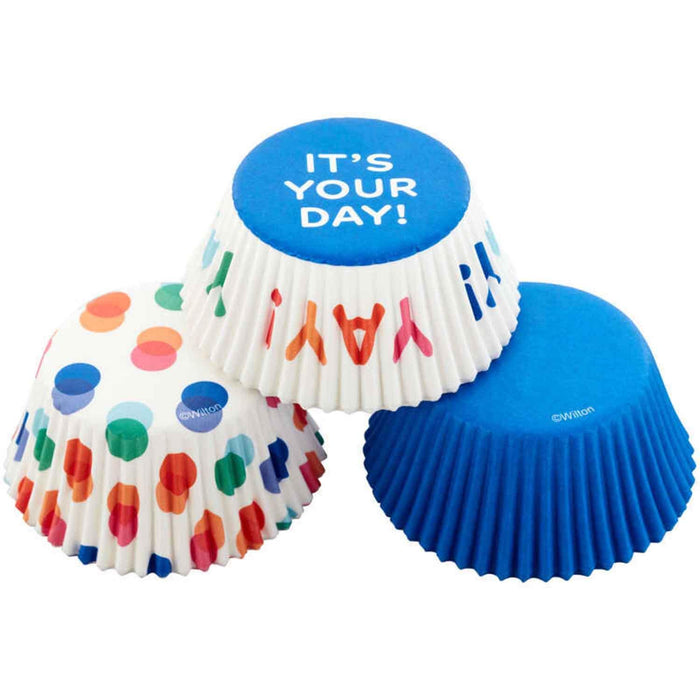 Three decorative baking cups in white, white with colorful dots and dashes, and solid blue, with the center cup displaying "IT'S YOUR DAY" on its blue top.
