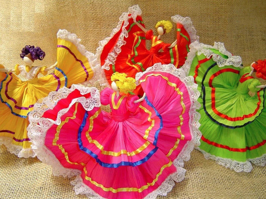 Four handmade corn husk dolls with colorful pleated fabric skirts in yellow, red, pink, and green, decorated with white lace trim and contrasting ribbon accents, displayed on a tan woven background.