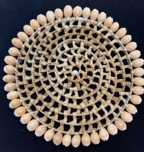 Circular placemat with cream-colored cowrie shells arranged around the border and woven tan geometric lattice pattern filling the center on a dark background.