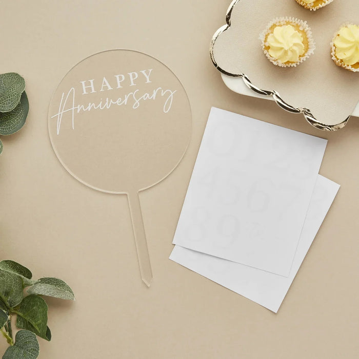Clear acrylic round cake topper with "HAPPY Anniversary" in white script text on a stick, displayed with blank white cards, potted succulent, and yellow frosted cupcakes on a beige background.
