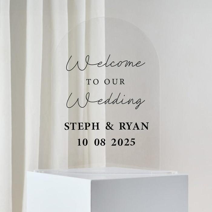 Clear acrylic arch-shaped wedding welcome sign with script and serif text reading "Welcome to our Wedding" and "Steph & Ryan 10 08 2025" displayed on a white pedestal base.