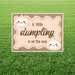 Rectangular yard sign with a warm beige background and wavy border, featuring cute smiling dumpling illustrations and the phrase “A little dumpling is on the way” in playful script, displayed on a lawn with a metal H-stake.