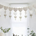 Beige paper heart garland with cascading nested hearts in graduating sizes, hung above a window with green potted plants below.