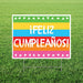 Colorful yard sign with horizontal stripes in green, yellow, blue, red, and orange displaying "¡FELIZ CUMPLEAÑOS!" in white text, decorated with white triangular bunting on top and bottom borders.