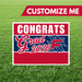 Red and navy blue graduation yard sign with "Congrats Grad 2026" text and customizable name area, displayed on grass with metal stake.