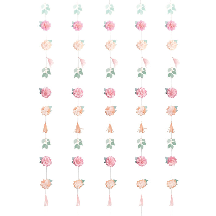 Five vertical strands of hanging paper flowers in pink and peach with green leaves and tassels arranged in a repeating pattern on a white background.