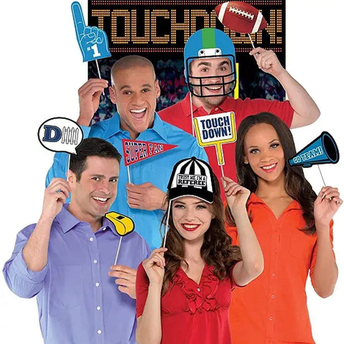 Five smiling people holding colorful football-themed photo booth props including a foam finger, helmet, referee whistle, touchdown sign, and megaphone against a scoreboard background.