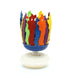 Colorful musical birthday candle with flame-shaped petals in red, blue, orange, green, yellow, and purple arranged in a circle on a blue base with white platform.