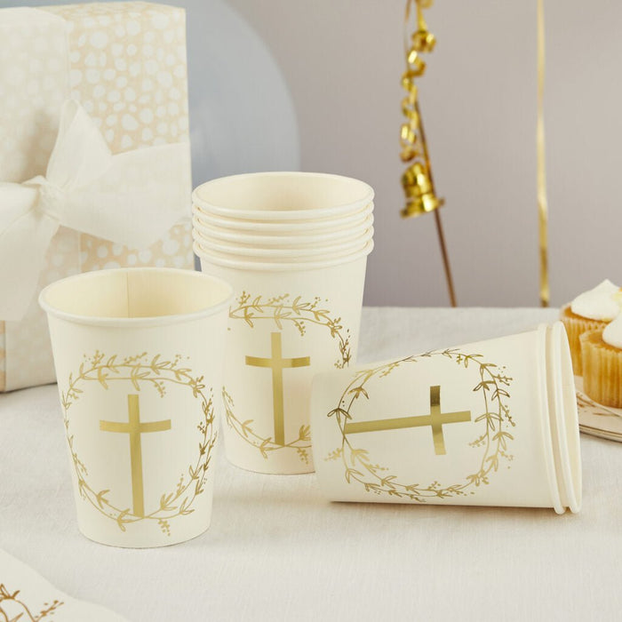 Eight cream-colored paper cups with gold cross and leaf wreath designs arranged on a white surface with matching napkins and decorative gold elements in the background.