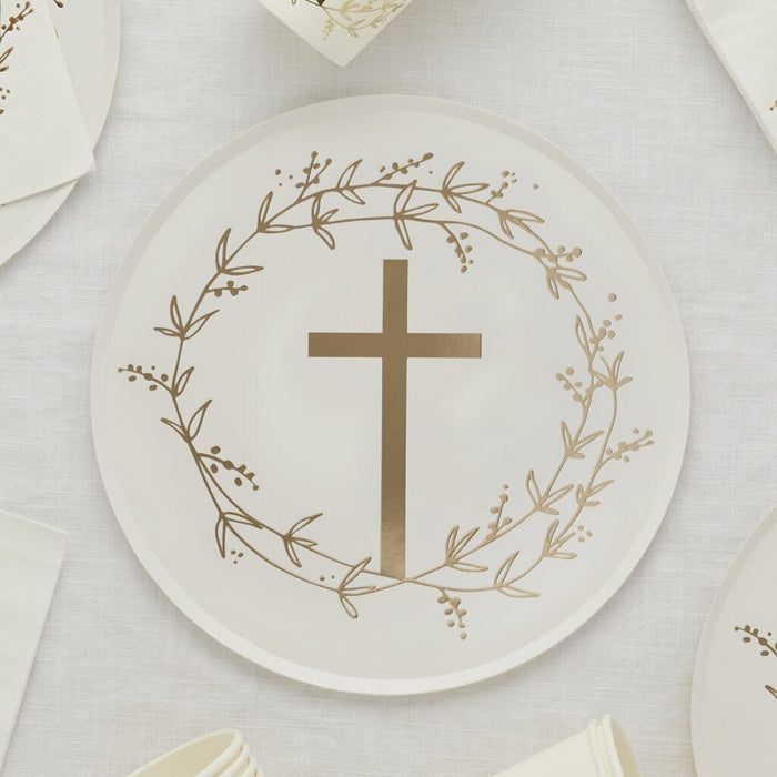 White paper plate with a gold cross at center surrounded by a delicate botanical wreath in gold, displayed on a cream linen surface with matching plates and napkins.