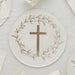 White paper plate with a gold cross at center surrounded by a delicate botanical wreath in gold, displayed on a cream linen surface with matching plates and napkins.