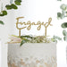 Gold script "Engaged" cake topper inserted into a white frosted cake decorated with baby's breath flowers, greenery, and a blush pink flower.