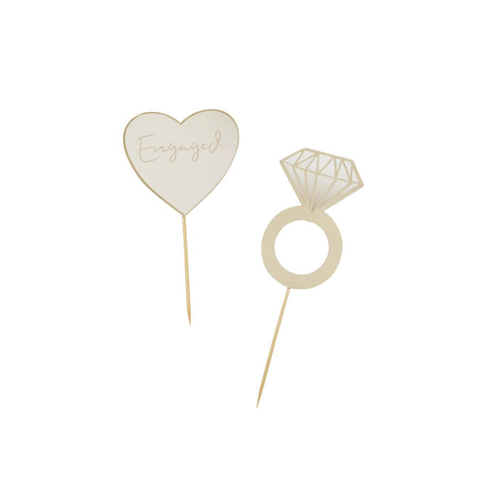 Two gold-colored wooden cake toppers on white background: a heart-shaped pick labeled "Engaged" and a diamond ring-shaped pick with geometric line detailing.