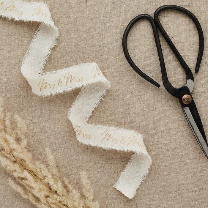 Cream-colored frayed cotton ribbon with gold "Mr & Mrs" text, displayed flat next to black scissors with wooden accents and dried pampas grass on beige linen.
