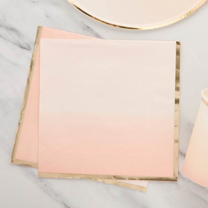 Stack of cream and pink ombre paper napkins with gold foil edges arranged on a marble surface with a gold-rimmed plate above.