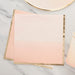Stack of cream and pink ombre paper napkins with gold foil edges arranged on a marble surface with a gold-rimmed plate above.