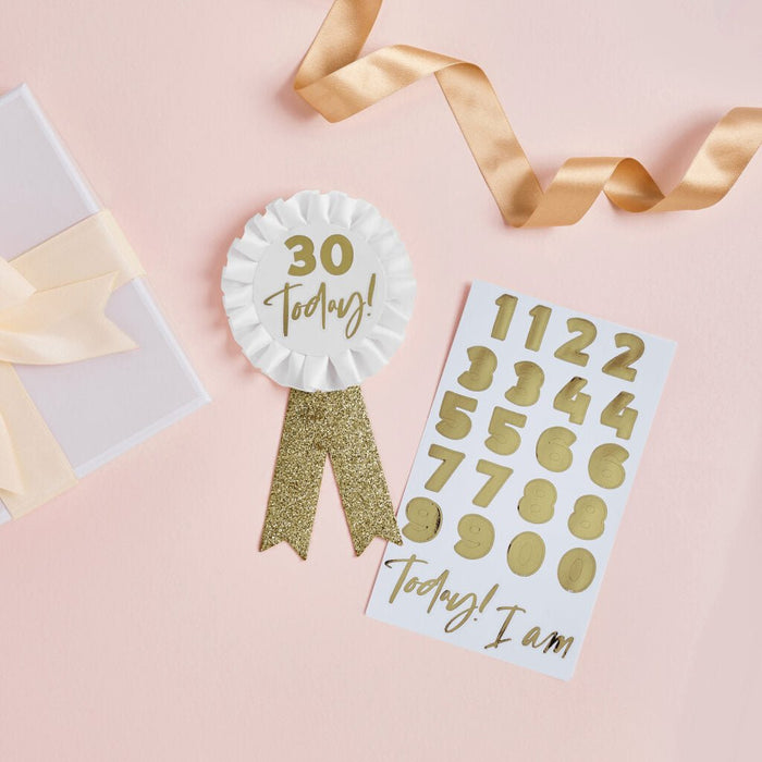 White rosette badge with gold "30 Today" text and glitter ribbon, displayed with a matching white card showing gold numbers and "Today I am" text, gold ribbons, and cream stationery on pink background.