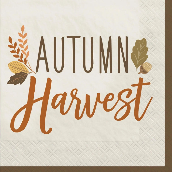 White beverage napkin with "Autumn Harvest" text in orange and brown lettering, decorated with autumn leaves and acorns in shades of orange, gold, and brown.