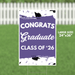Purple and white yard sign reading "Congrats Graduate Class of '26" with black graduation cap decorations and confetti motifs throughout the design.