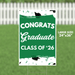 Large green yard sign with white text reading "CONGRATS Graduate CLASS OF '26" decorated with black graduation caps and green confetti, measuring 24 inches by 36 inches.
