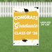 Large yellow yard sign with white text reading "Congrats Graduate Class of '26" decorated with black graduation caps and gold confetti designs, measuring 24 by 36 inches.