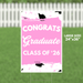 Pink yard sign with white text reading "CONGRATS Graduate CLASS OF '26" decorated with black graduation caps and pink confetti designs.