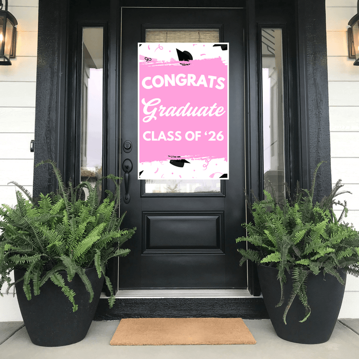 Hot pink graduation door banner with white "CONGRATS Graduate CLASS OF '26" text and black mortarboard accents, displayed on a black front door flanked by fern planters.