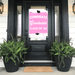 Hot pink graduation door banner with white "CONGRATS Graduate CLASS OF '26" text and black mortarboard accents, displayed on a black front door flanked by fern planters.