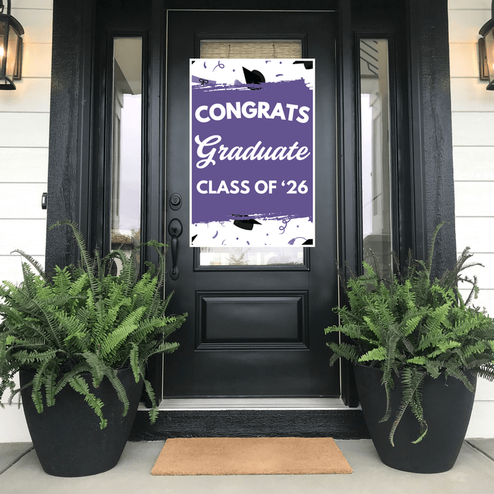 Purple and white graduation door banner reading "CONGRATS Graduate CLASS OF '26" with black mortarboard icons, displayed on a black front door flanked by fern planters.