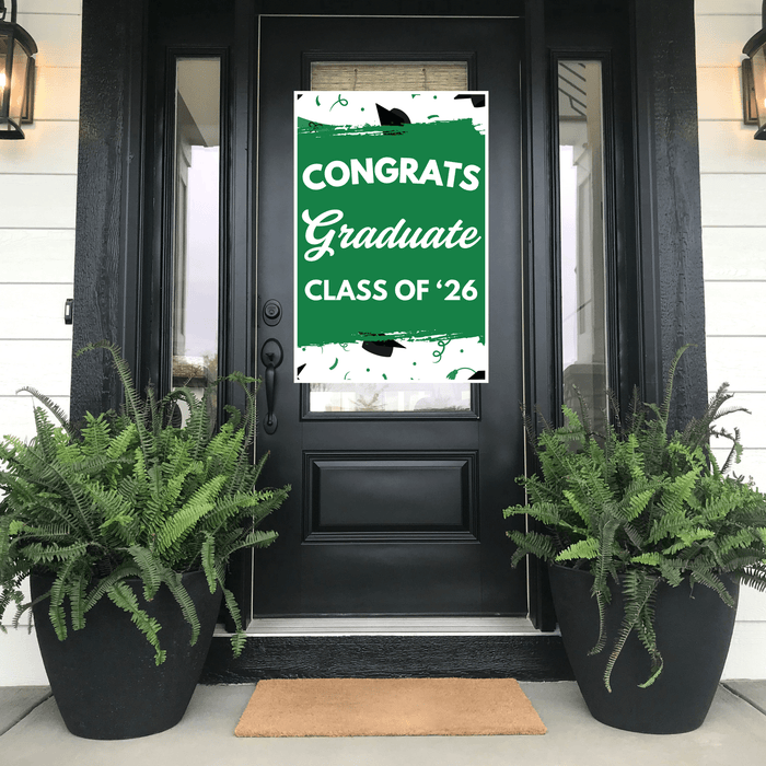 Green and white graduation door banner reading "CONGRATS Graduate CLASS OF '26" with mortarboard graphics, displayed on a black front door flanked by fern planters.