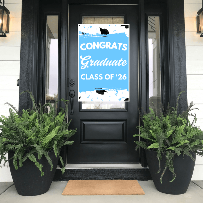 Light blue graduation door banner reading "CONGRATS Graduate CLASS OF '26" with black cap and tassel illustrations, displayed on a black front door flanked by fern planters.
