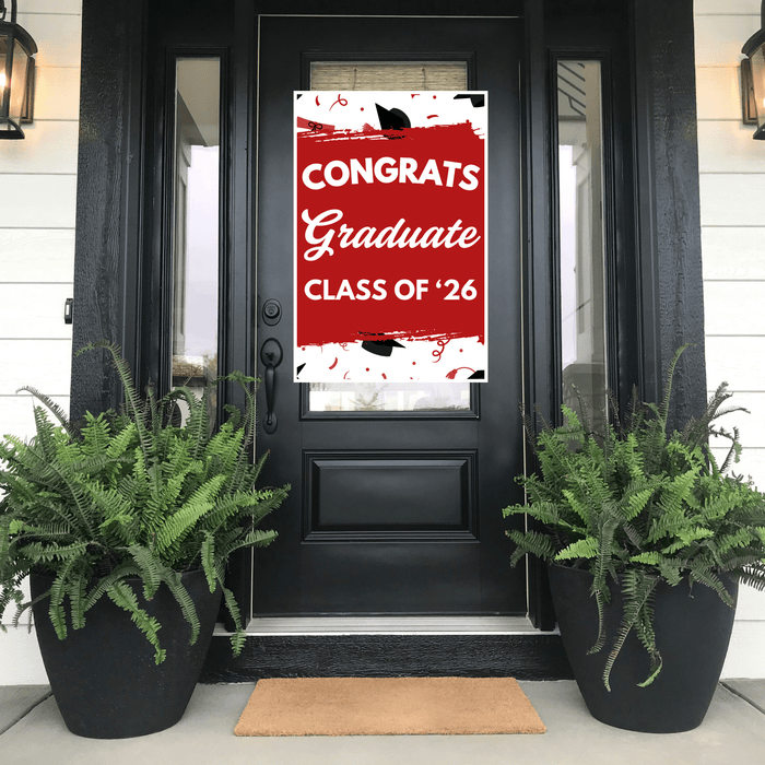 Red and white "Congrats Graduate Class of '26" door banner displayed on a black front door with glass panels, flanked by two black planters with green ferns.