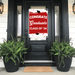 Red and white "Congrats Graduate Class of '26" door banner displayed on a black front door with glass panels, flanked by two black planters with green ferns.