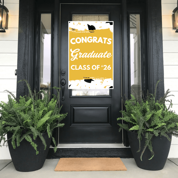 Yellow and white graduation door banner reading "Congrats Graduate Class of '26" with black mortarboard graphics, displayed on a black front door flanked by fern planters.